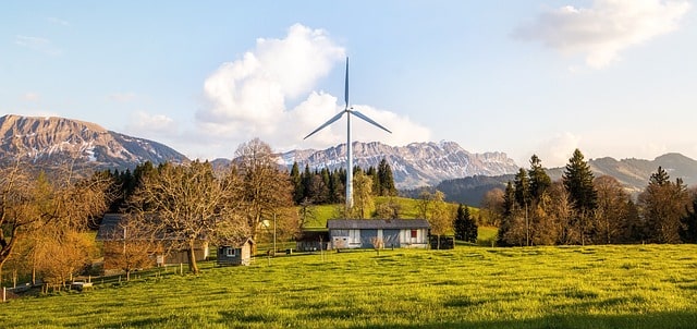 Bild på ett vindkraftverk bredvid ett hus som visar hur stort ett vindkraftverk verkligen är, med ett bergsmassiv i bakgrunden.